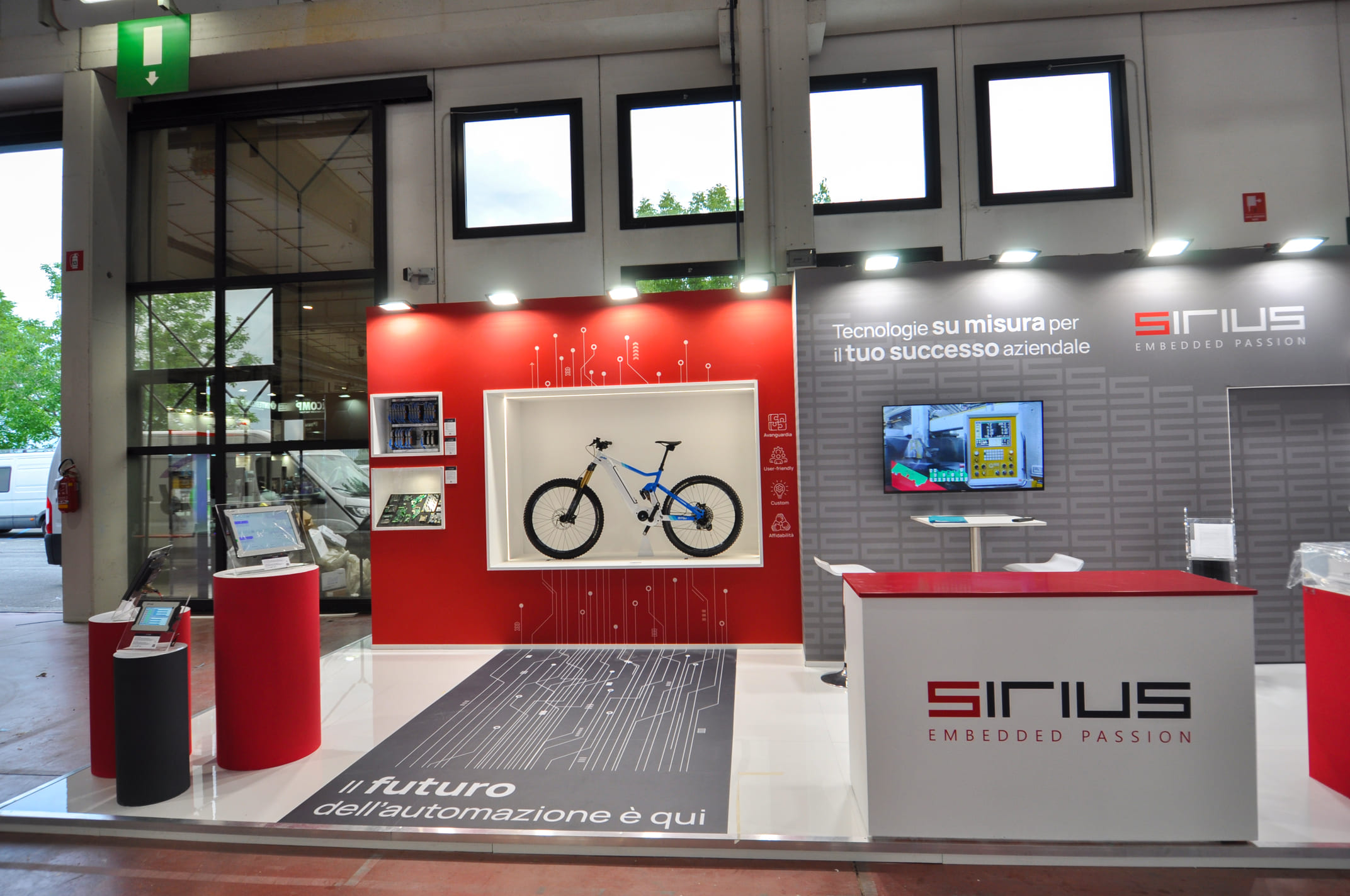 Fronte dello stand Sirius con nicchia LED, tablet su cilindri espositivi e bancone con logo
