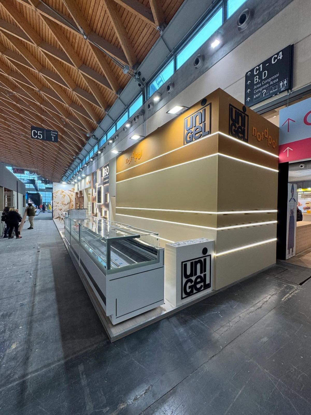 Angolo dello stand visto dalla corsia: vetrina espositiva e pareti crema con strisce luminose e loghi Unigel e Nordpol